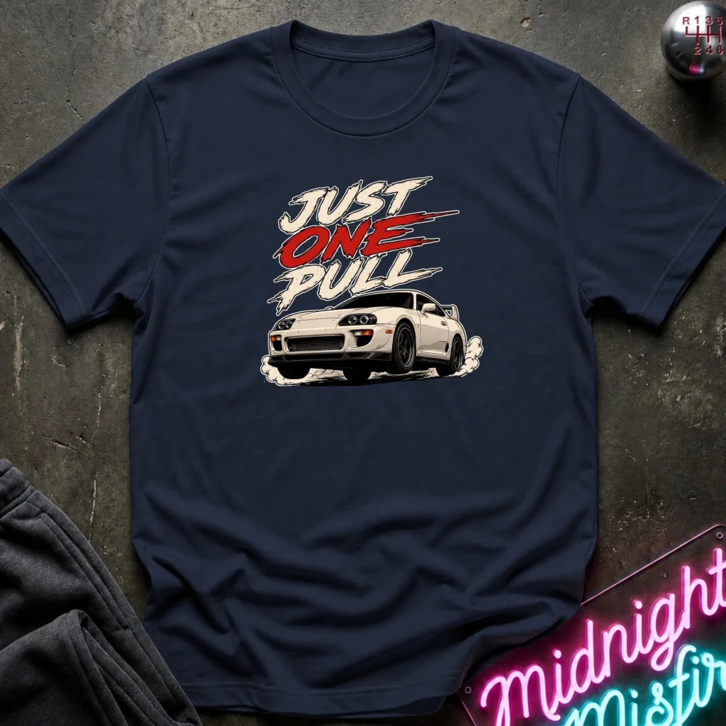 A navy blue graphic t-shirt laid flat on a dark textured surface. The shirt features a white sports car and the text 'JUST ONE PULL' in white and red.