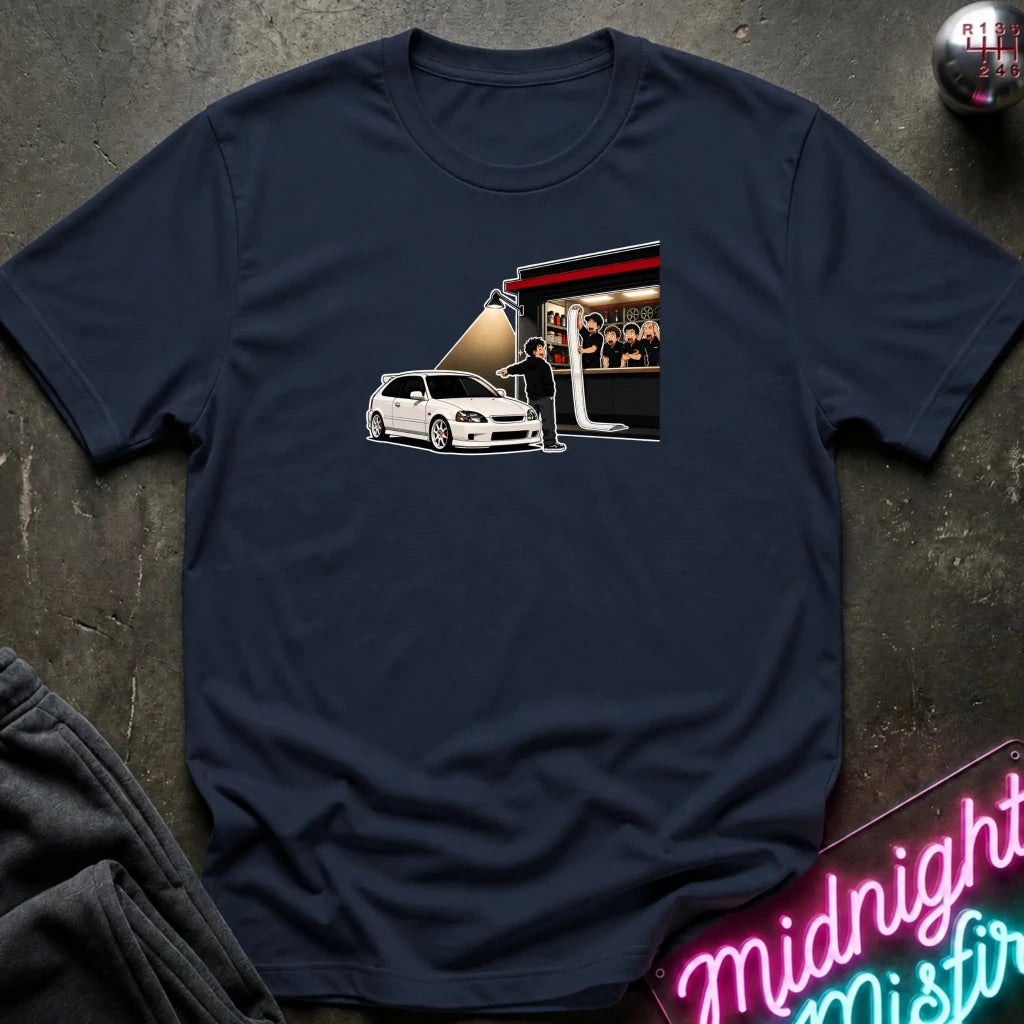 A navy blue crewneck t-shirt laid flat on a dark textured surface, featuring a graphic print of a white hatchback car next to a stage with people. 