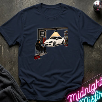 A navy blue short-sleeve t-shirt with a graphic print of a person sitting in a garage chair looking at a white sports car outside an open garage. The shirt is displayed flat on a dark textured surface with a neon sign and sweatpants partially visible.