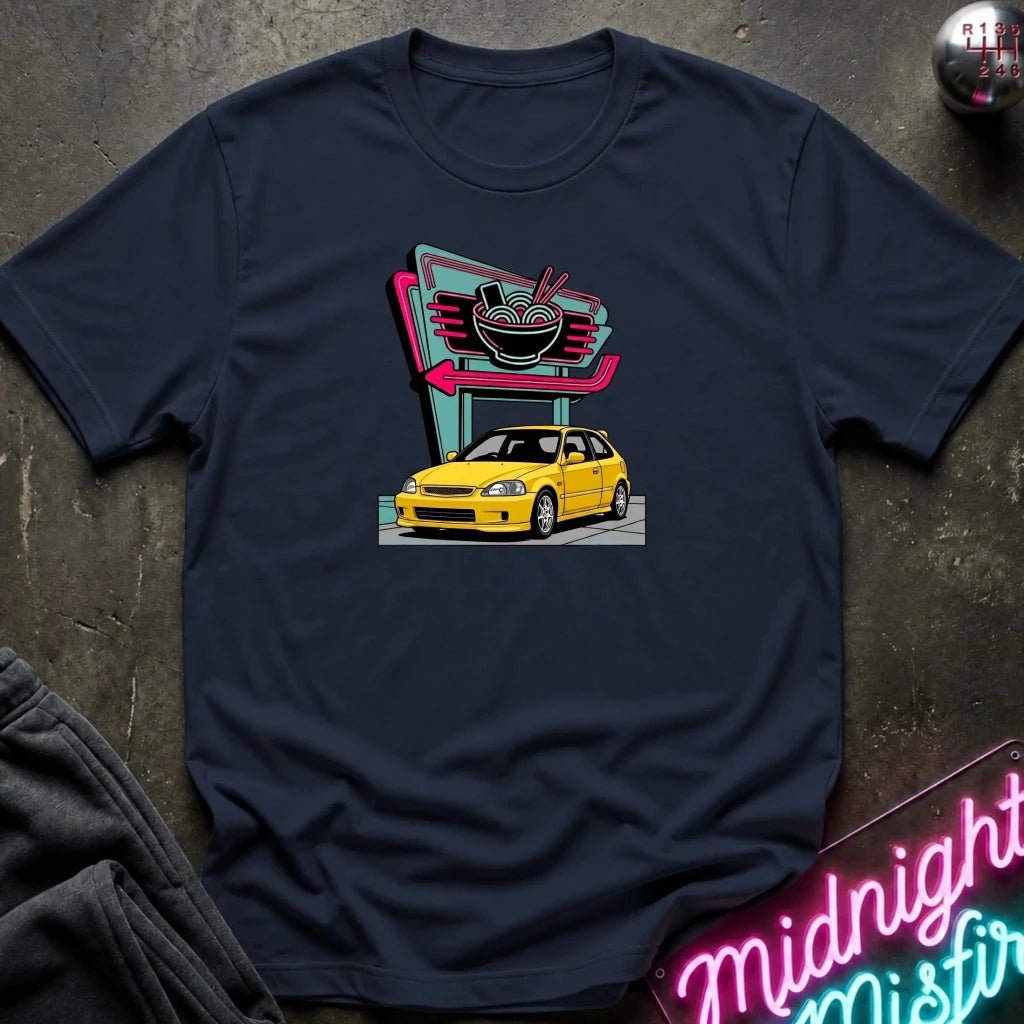 A navy blue short-sleeve t-shirt laid flat on a dark textured surface, featuring a retro-style graphic of a yellow hatchback car parked in front of a neon ramen shop sign. 