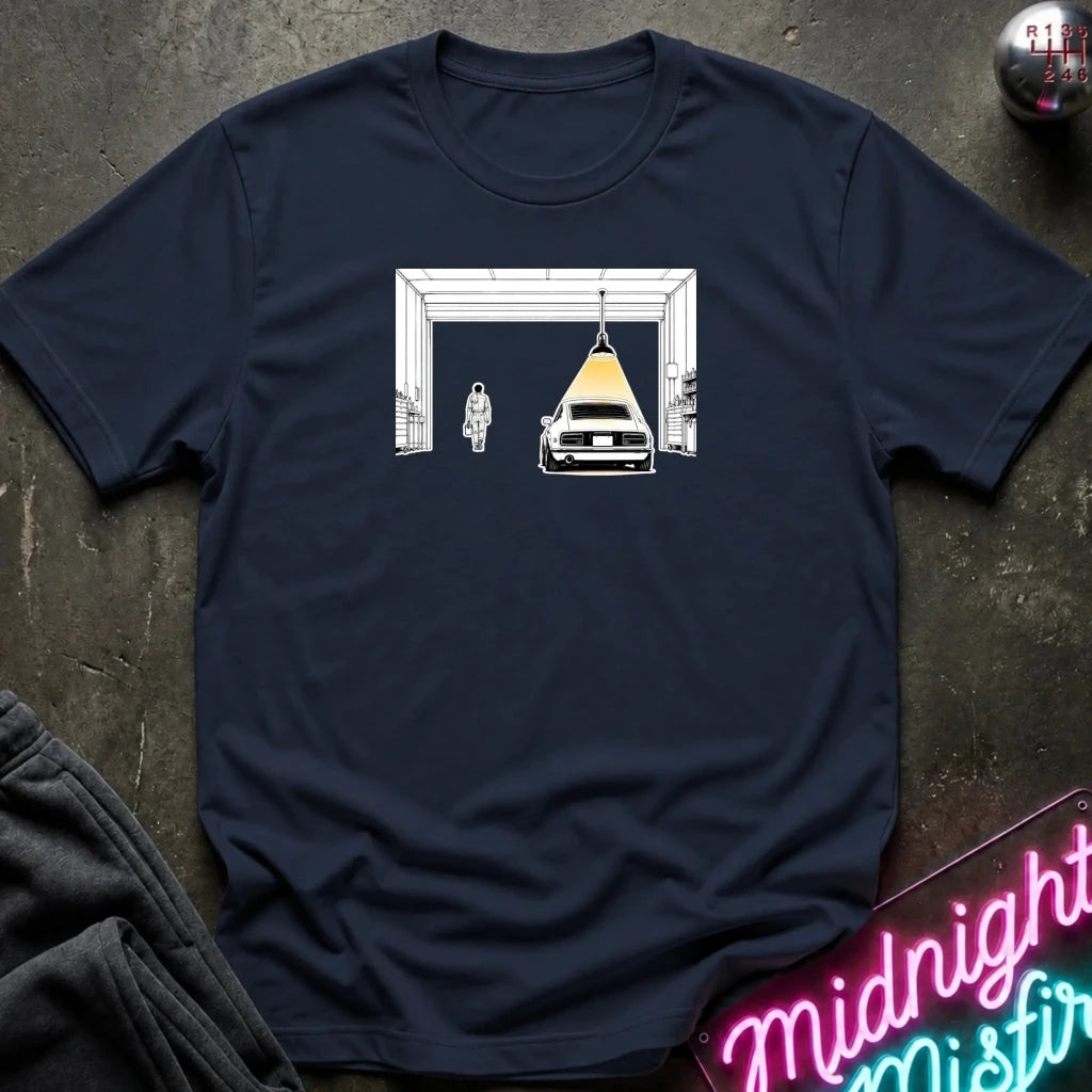 A navy blue short-sleeve t-shirt laid flat on a dark textured surface, featuring a graphic print of a white car under a yellow tent suspended by ropes, with a white archway behind it. A folded dark gray garment and a neon sign are partially visible at the edges.