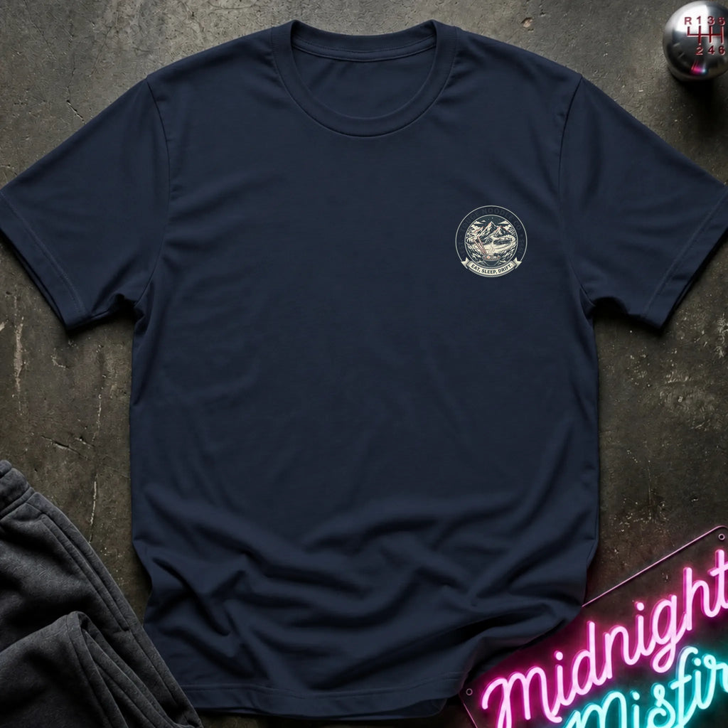 Navy crewneck t-shirt with a circular crest printed on the left chest, styled flat on a dark textured surface with a neon sign and dark pants in the background.