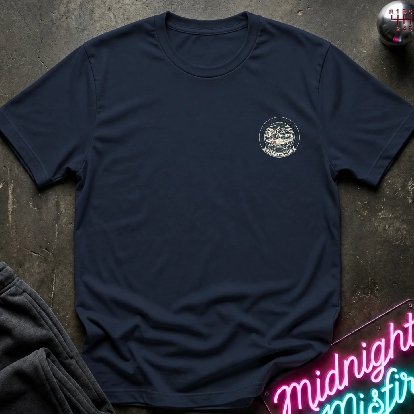 Navy crewneck t-shirt with a circular crest printed on the left chest, styled flat on a dark textured surface with a neon sign and dark pants in the background.