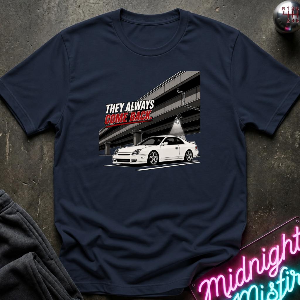 A navy blue graphic t-shirt laid flat on a dark textured surface. The graphic features a white sports car parked under a highway overpass with the text 'THEY ALWAYS COME BACK' in white and red.