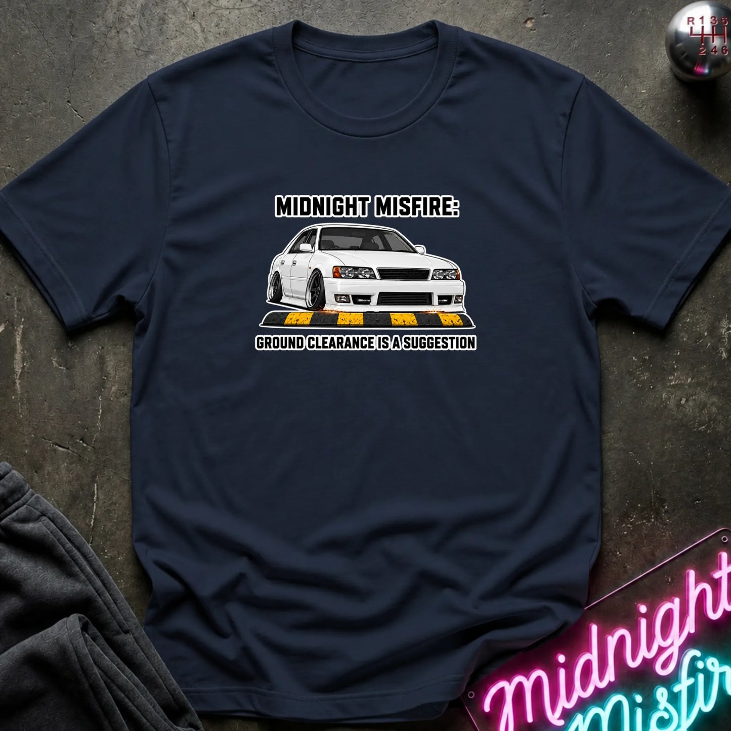 A navy blue graphic t-shirt laid flat on a dark textured surface. The shirt features a white car graphic with the text 'MIDNIGHT MISFIRE: GROUND CLEARANCE IS A SUGGESTION'.