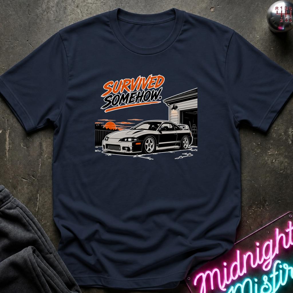 A navy blue graphic t-shirt laid flat on a dark textured surface. The shirt features a graphic of a black sports car in front of a garage with the text 'SURVIVED SOMEHOW!' in orange and white.