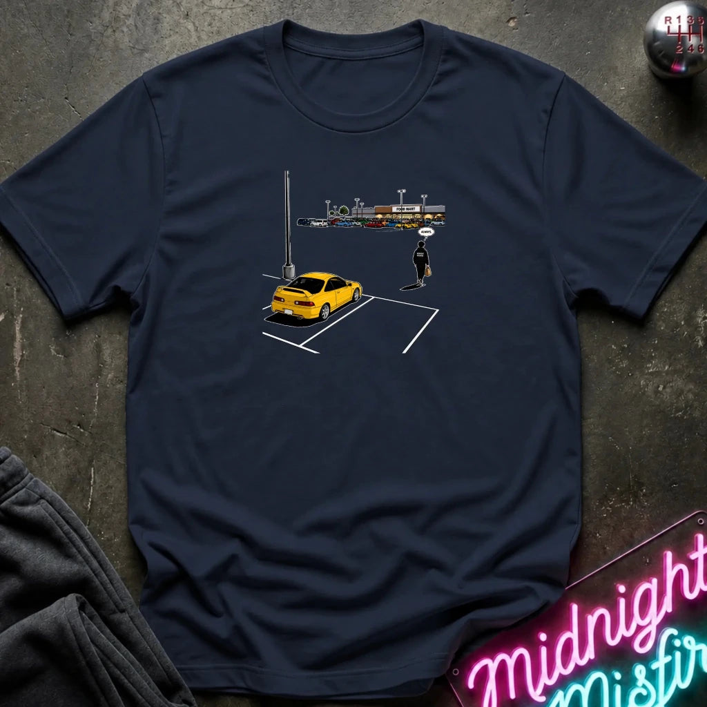 A navy blue short-sleeve t-shirt with a graphic print of a yellow sports car parked in a spot, laid flat on a dark textured surface with a neon sign and sweatpants partially visible.