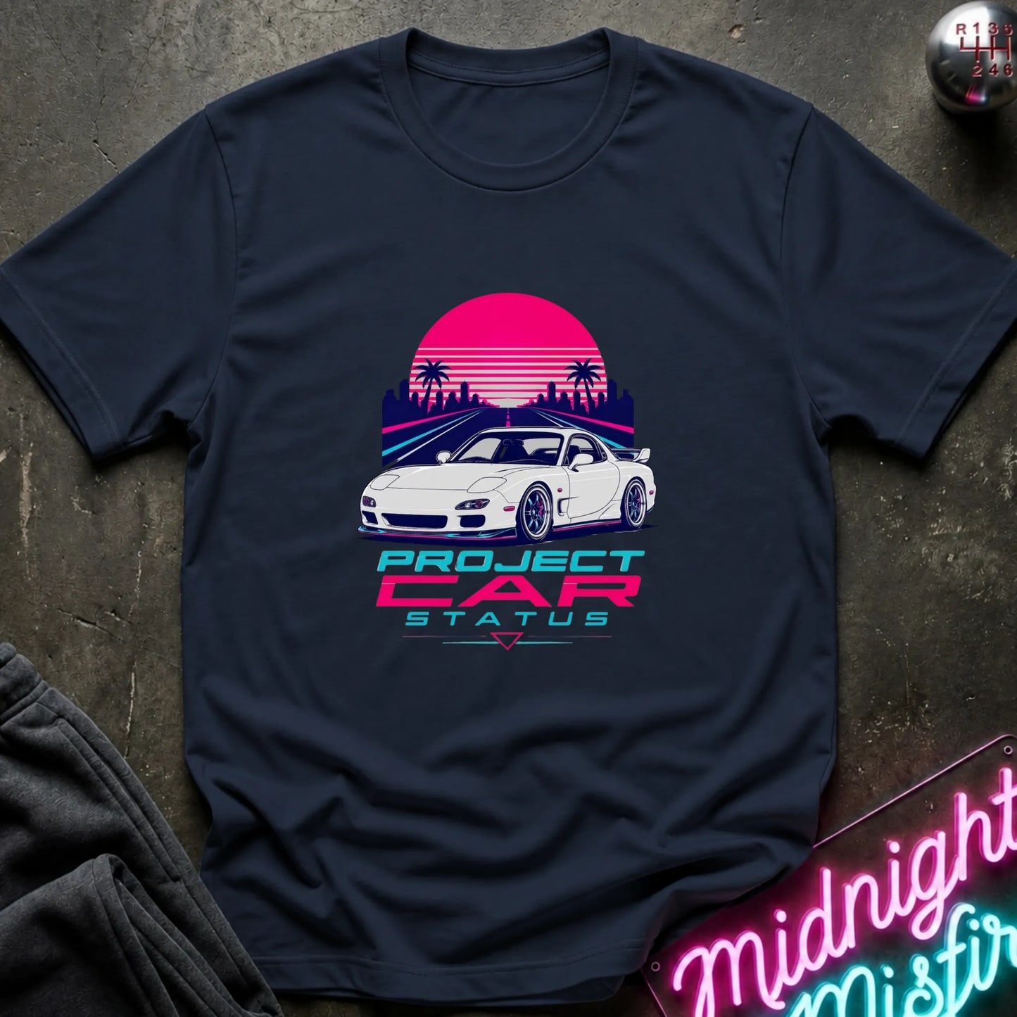 A navy short-sleeve graphic T-shirt laid flat on a dark textured surface. The shirt features a retro-style print of a white sports car in front of a pink and purple sunset with palm trees, and the text 'PROJECT CAR STATUS' below.