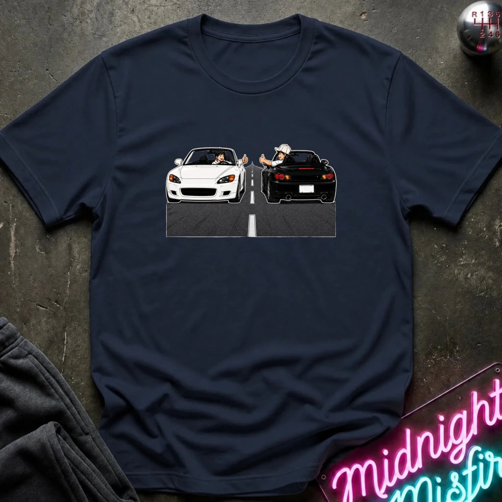 A navy blue short-sleeve t-shirt laid flat on a dark textured surface, featuring a graphic print of a white convertible and a black sports car facing each other on a road with a cartoon character in the middle. A neon sign and folded dark fabric are partially visible in the corners.