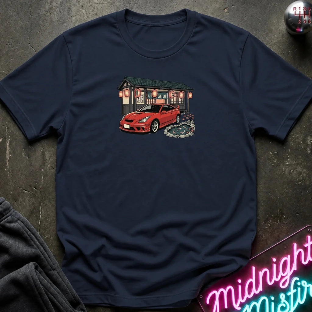 A navy blue short-sleeve t-shirt laid flat on a dark textured surface, featuring a graphic print of a red sports car parked in front of a building with glowing lanterns. 