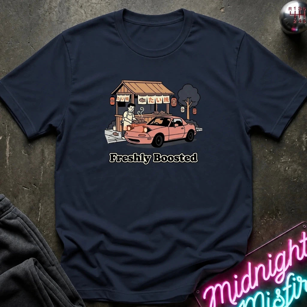 A navy blue short-sleeve t-shirt laid flat on a dark textured surface, featuring a graphic of a pink sports car parked in front of a small food stall, with the text 'Freshly Boosted' printed below.