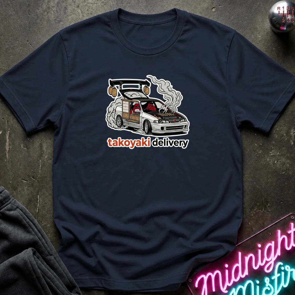 A navy blue short-sleeve t-shirt laid flat on a dark textured surface, featuring a graphic of a white car with smoke and the text 'takoyaki delivery' in orange and white. 
