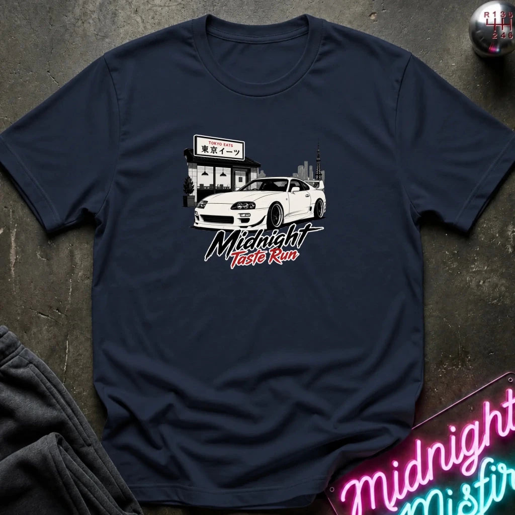 A navy blue short-sleeve t-shirt laid flat on a dark textured surface, featuring a graphic print of a white sports car in front of a building with Japanese text, and the words 'Midnight Taste Run' in stylized white and red lettering. 