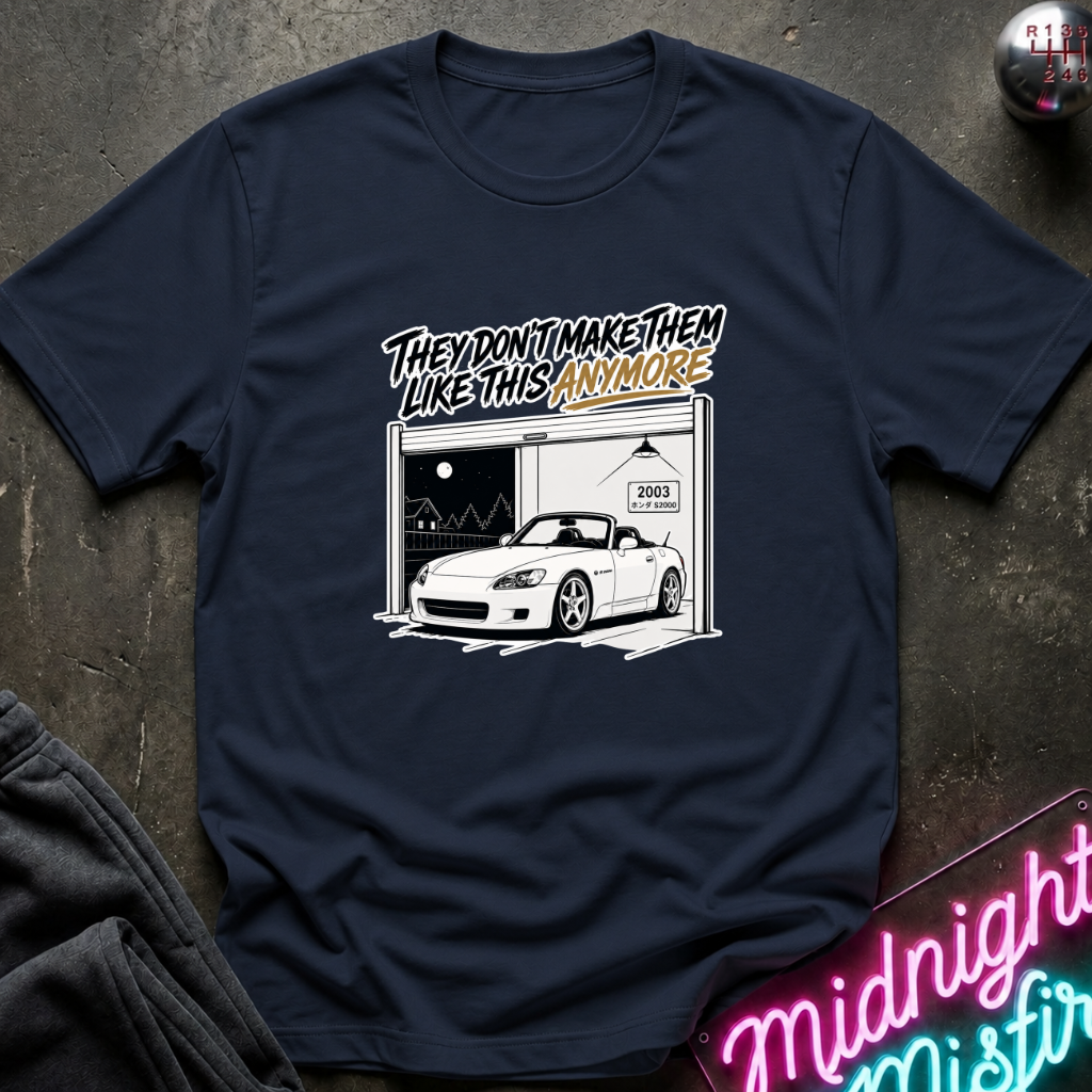 A navy blue graphic t-shirt laid flat on a dark textured surface. The graphic features a white convertible sports car parked in front of a garage with the text 'THEY DON'T MAKE THEM LIKE THIS ANYMORE' in stylized lettering.