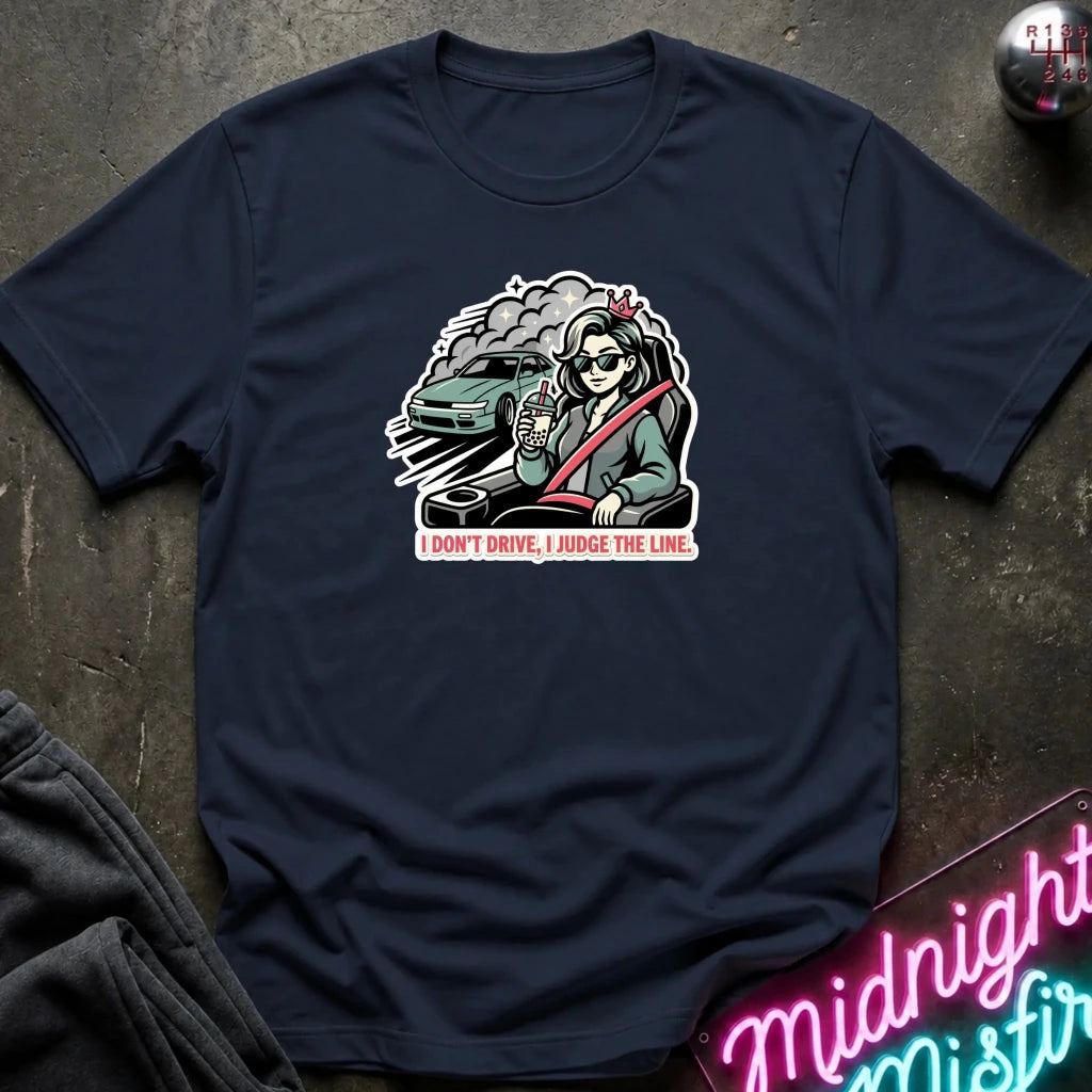 A navy blue short-sleeve t-shirt laid flat on a dark textured surface, featuring a central graphic of a skeleton driver wearing a seatbelt next to a green car with the text 'I DON'T DRIVE, I JUDGE THE LINE.' A neon sign and folded dark fabric are partially visible in the corners.