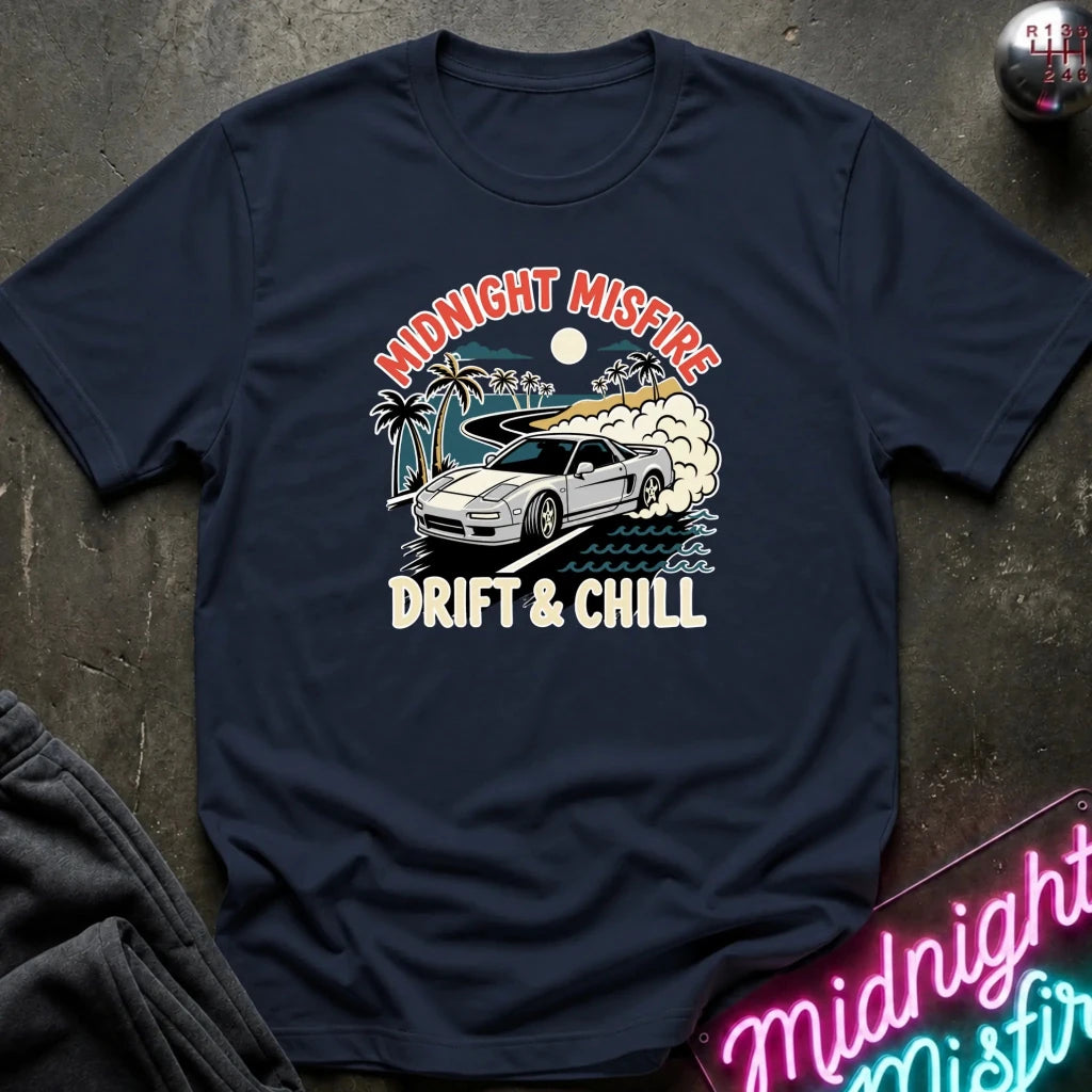 A navy blue graphic t-shirt laid flat on a dark textured surface. The shirt features a retro-style print of a white sports car drifting with palm trees, a surfboard, and the text 'MIDNIGHT MISFIRE' and 'DRIFT & CHILL'.