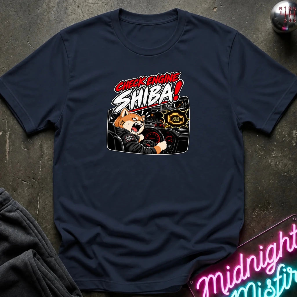 A navy blue graphic t-shirt laid flat on a dark textured surface. The shirt features a cartoon Shiba Inu driving a car with the text 'CHECK ENGINE SHIBA!' in red and white.