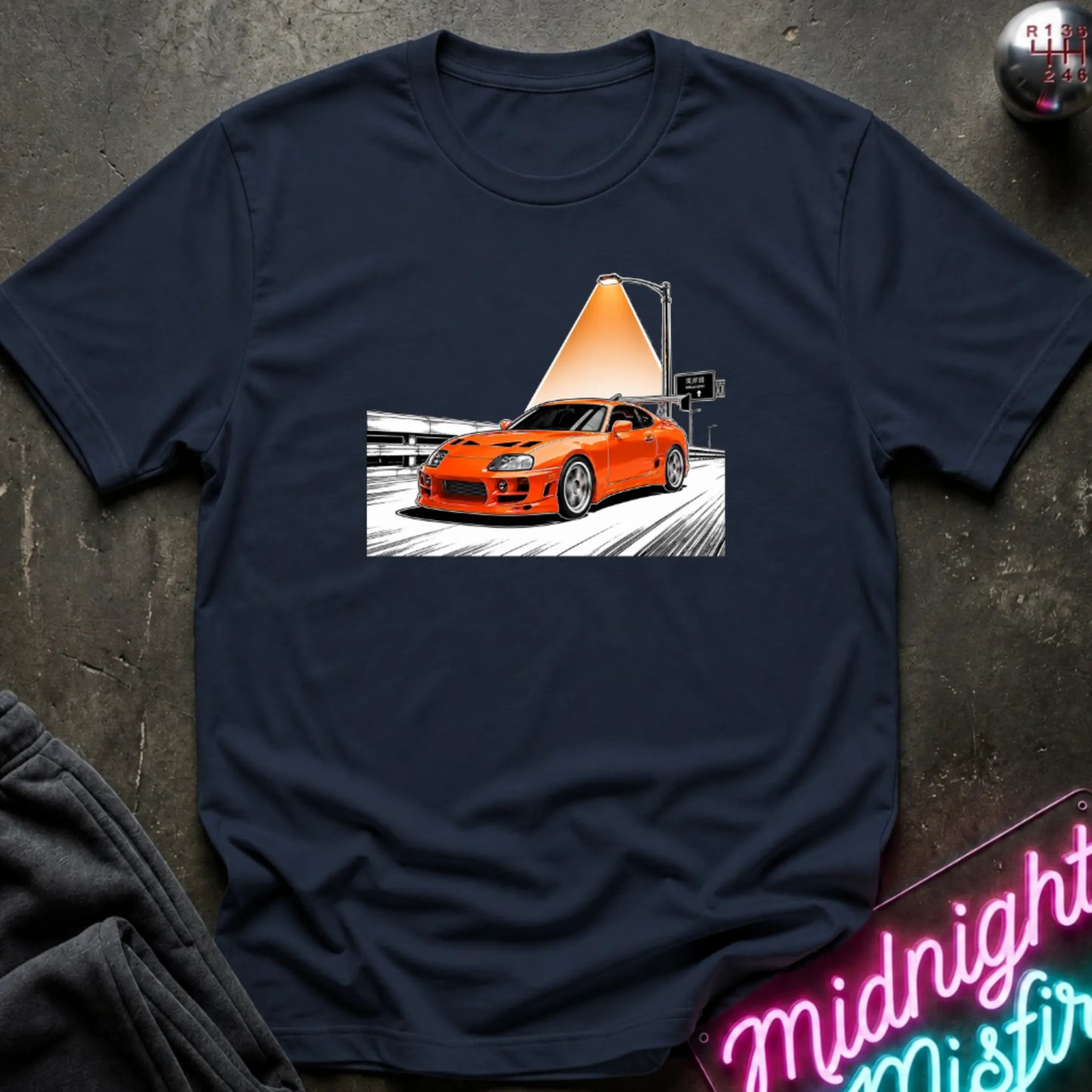 A navy short-sleeve t-shirt laid flat on a dark textured surface, featuring a graphic print of an orange sports car speeding on highway.