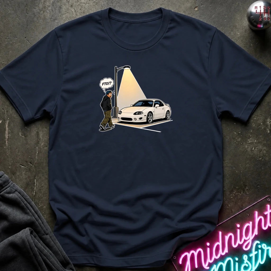A navy blue graphic t-shirt laid flat on a dark textured surface. The graphic shows a white sports car parked under a streetlamp with a cartoon figure and a speech bubble 
