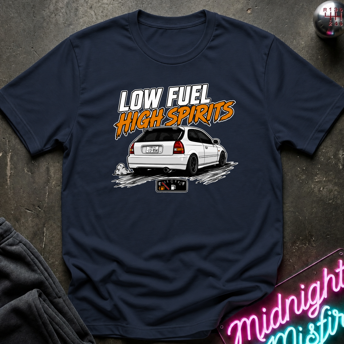 A navy blue graphic t-shirt laid flat on a dark textured surface. The shirt features a white car graphic with the text 'LOW FUEL HIGH SPIRITS' in white and orange.