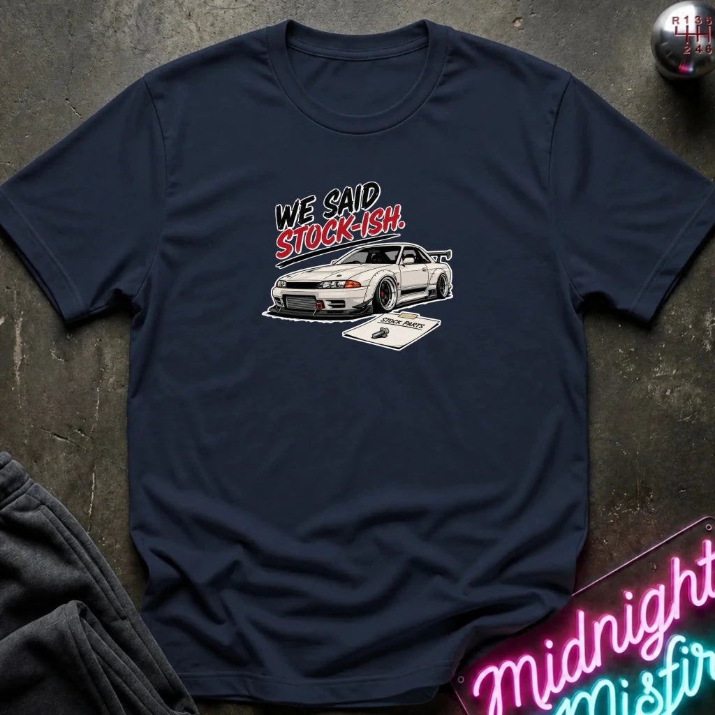 A navy blue graphic t-shirt laid flat on a dark textured surface. The shirt features a white sports car and the text 'WE SAID STOCK-ISH.