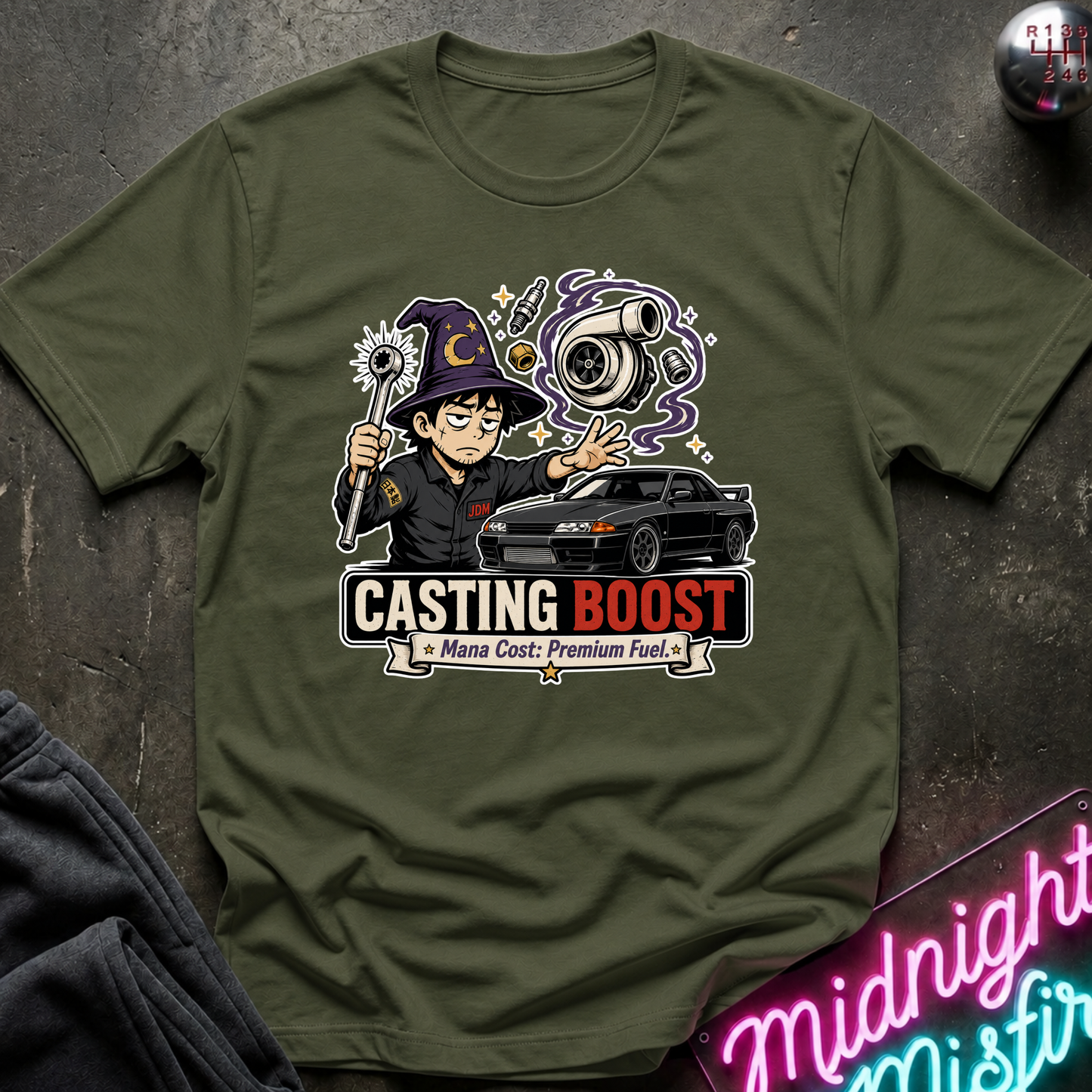 An olive green graphic t-shirt laid flat on a dark textured surface. The graphic features a cartoon wizard holding a wand next to a black sports car, with the text 'CASTING BOOST' and 'A Mana Cost: Premium Fuel'.