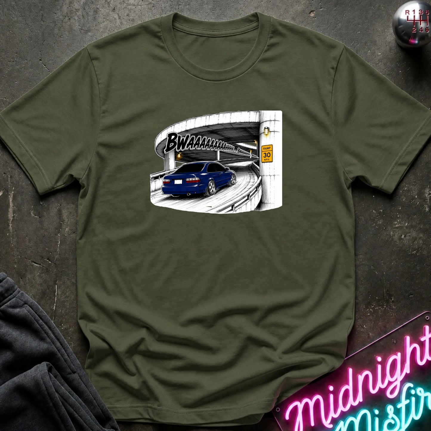 An olive green short-sleeve crewneck t-shirt laid flat on a dark textured surface. The shirt features a graphic print of a blue car driving under a highway overpass with the text 'BWAHAHA'.