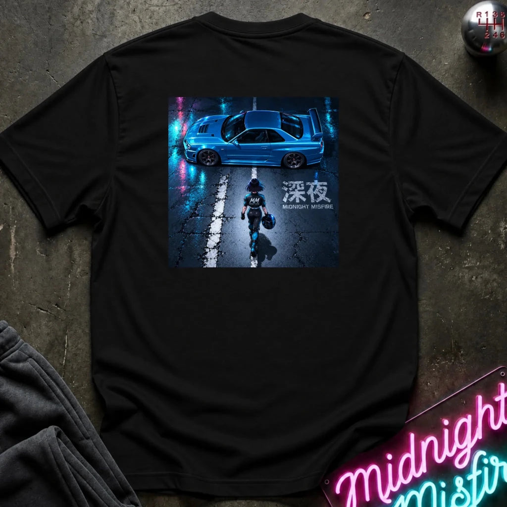 A black short-sleeve t-shirt laid flat on a dark textured surface, featuring a large rectangular graphic print on the back with a blue car, a dark figure, and neon-style text. A folded dark garment and a pink neon sign are partially visible at the edges as styling props.