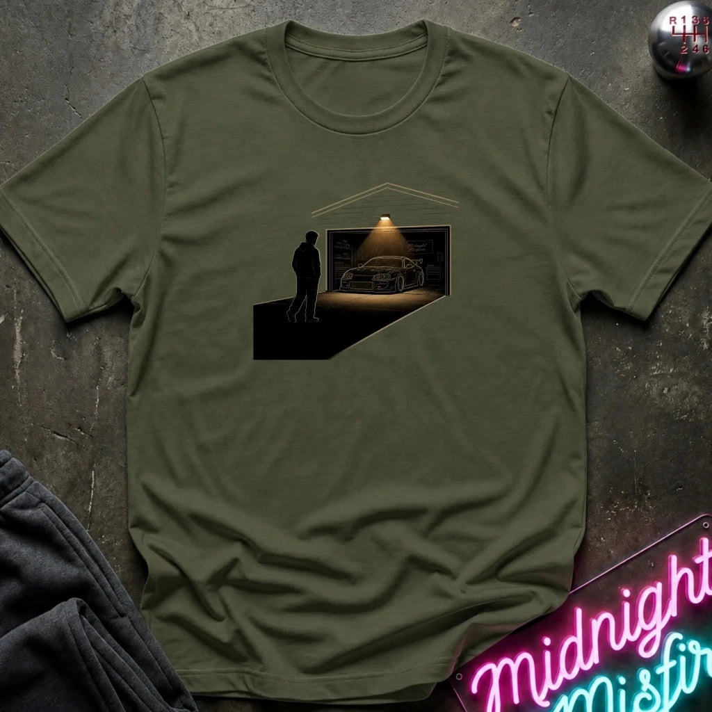An olive green crewneck t-shirt laid flat on a dark textured surface, featuring a graphic print of a silhouette of a man looking at a sports car inside a garage. 