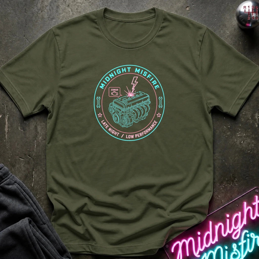 An olive green short-sleeve t-shirt with a teal and white graphic print reading 'MIDNIGHT MISFIRE' around an engine illustration, laid flat on a dark textured surface with a neon sign and dark pants partially visible.