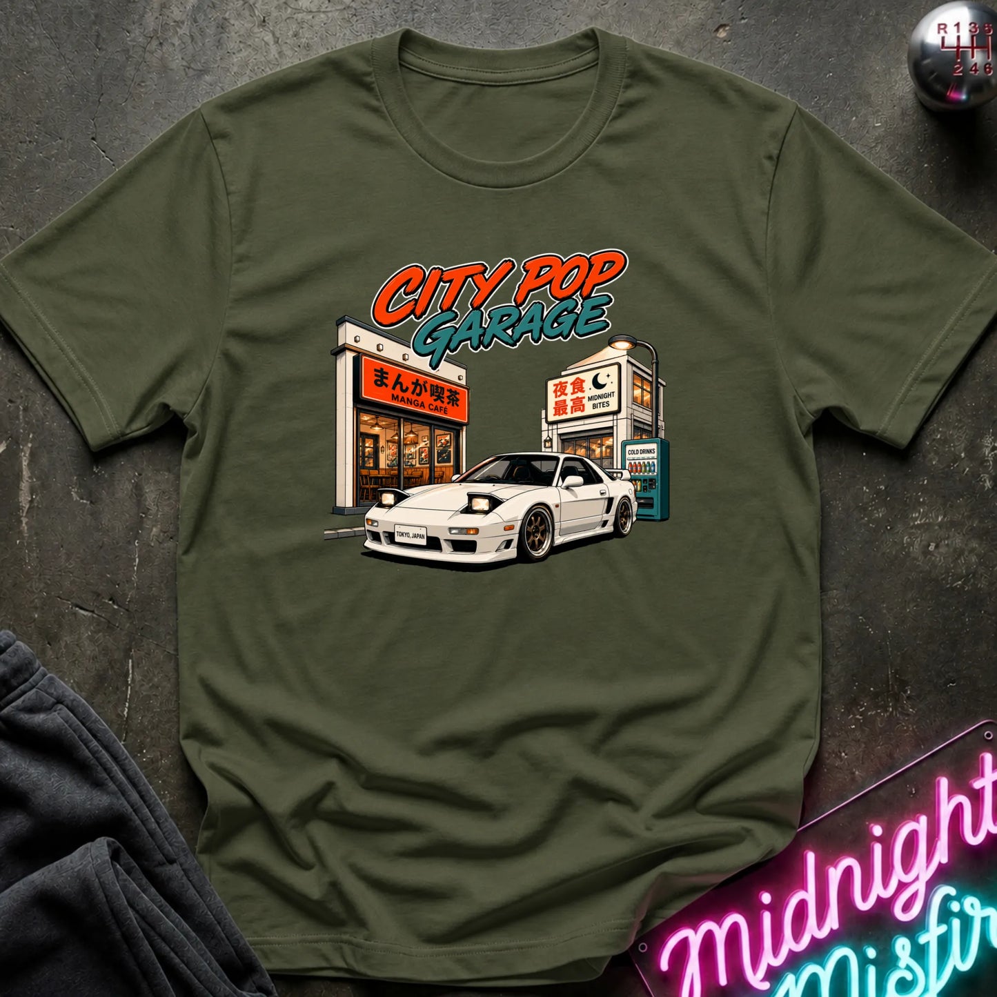 An olive green short-sleeve graphic t-shirt laid flat on a dark textured surface. The graphic features a white sports car parked in front of a cafe with the text 'CITY POP GARAGE' in orange and teal.