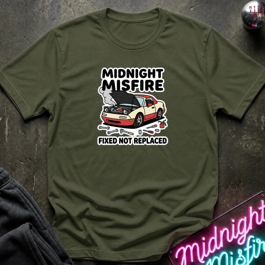 An olive green short-sleeve t-shirt with a graphic print of a car with its hood open, accompanied by the text 'MIDNIGHT MISFIRE' and 'FIXED NOT REPLACED'. The shirt is displayed flat on a dark textured surface with a neon sign and dark pants partially visible.