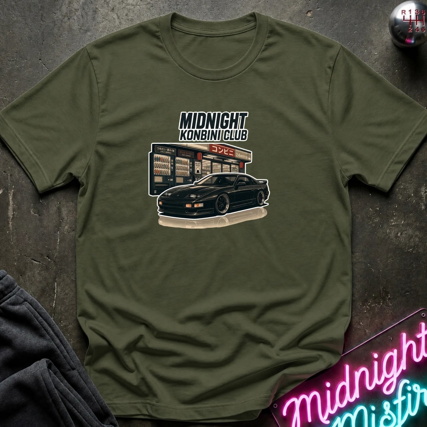 An olive green short-sleeve graphic t-shirt laid flat on a dark textured surface. The graphic features the text 'MIDNIGHT KONBINI CLUB' above an illustration of a black sports car parked in front of a convenience store.
