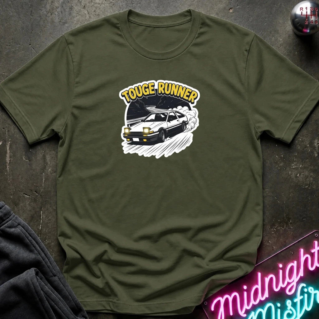 An olive green short-sleeve t-shirt with a 'TOUGE RUNNER' graphic featuring a black car and yellow text, laid flat on a dark textured surface with a neon sign and dark pants partially visible.