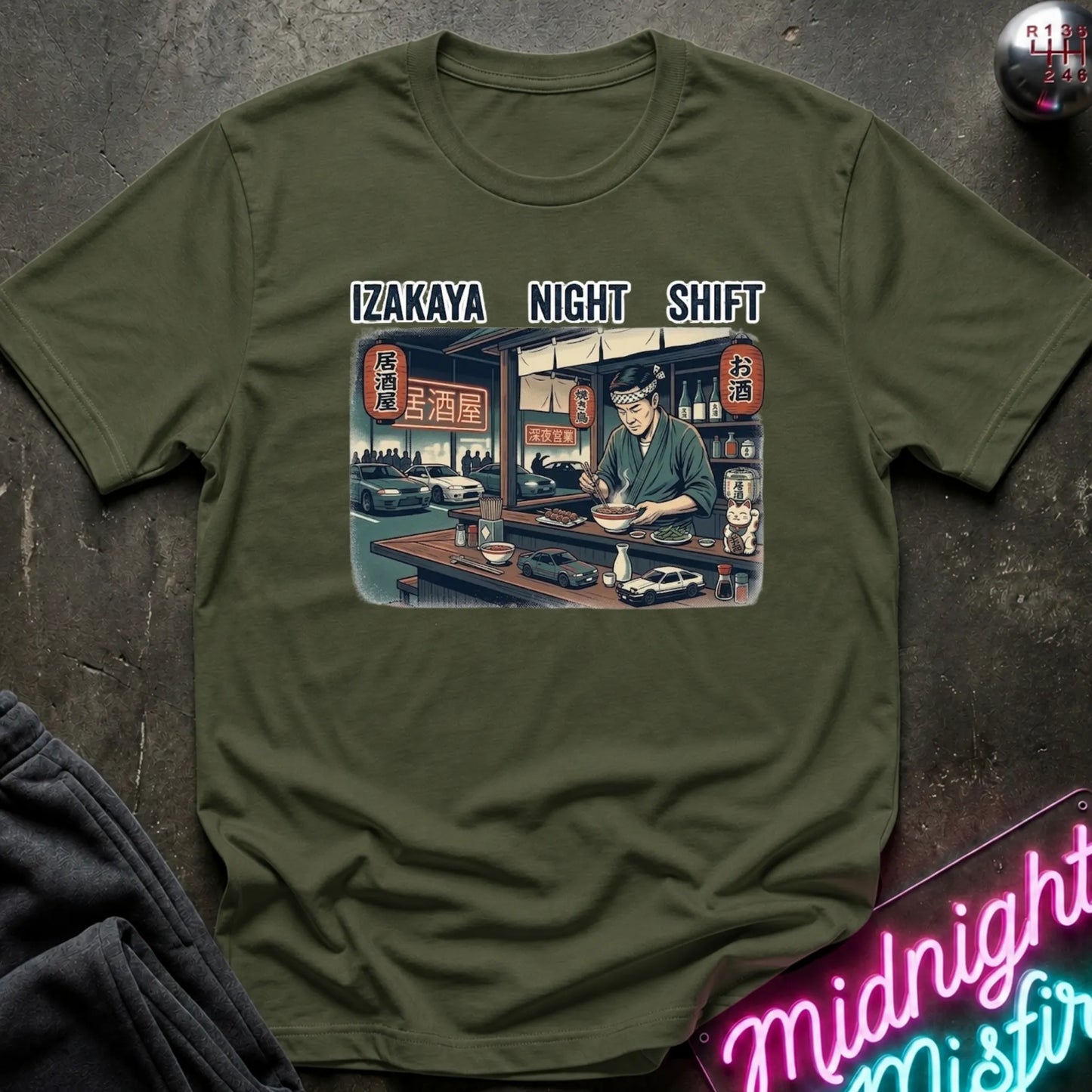 An olive green crewneck t-shirt featuring a graphic print of an Izakaya night shift bartender with the text 'IZAKAYA NIGHT SHIFT' above it, styled on a dark textured surface with a neon sign and dark jacket in the background.