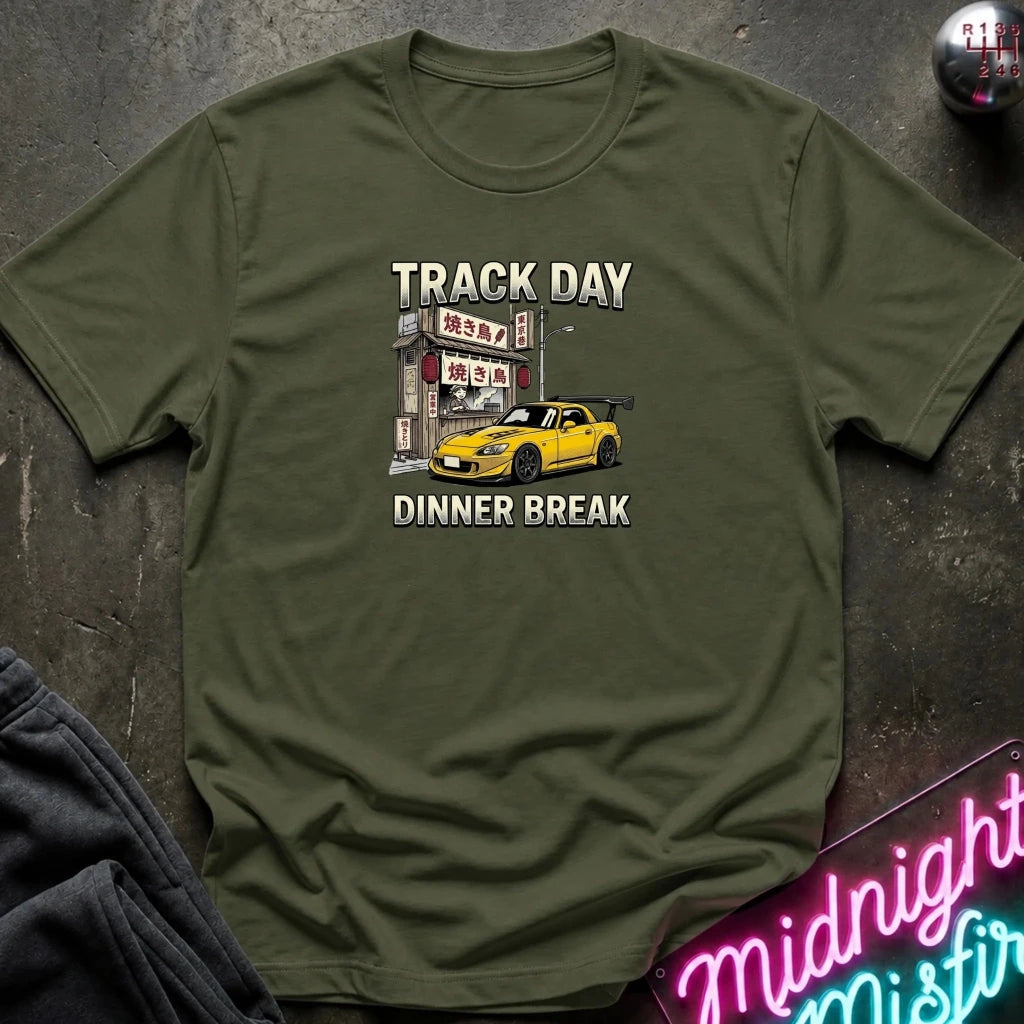 An olive green short-sleeve t-shirt with a graphic print of a yellow sports car parked in front of a Japanese street sign, with the text 'TRACK DAY DINNER BREAK' printed in white. The shirt is laid flat on a dark textured surface with a neon sign and dark pants partially visible.