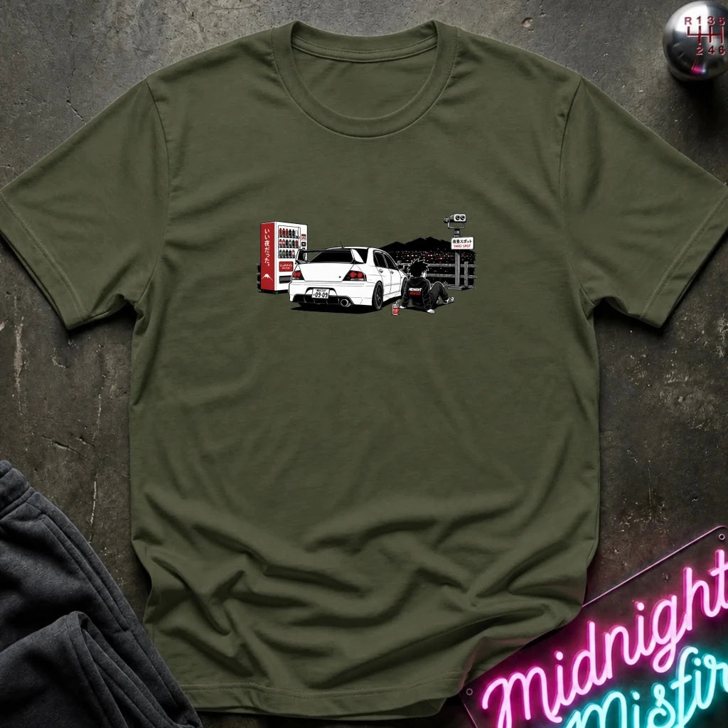 An olive green short-sleeve t-shirt laid flat on a dark textured surface, featuring a graphic print of a white sports car with a red vending machine.