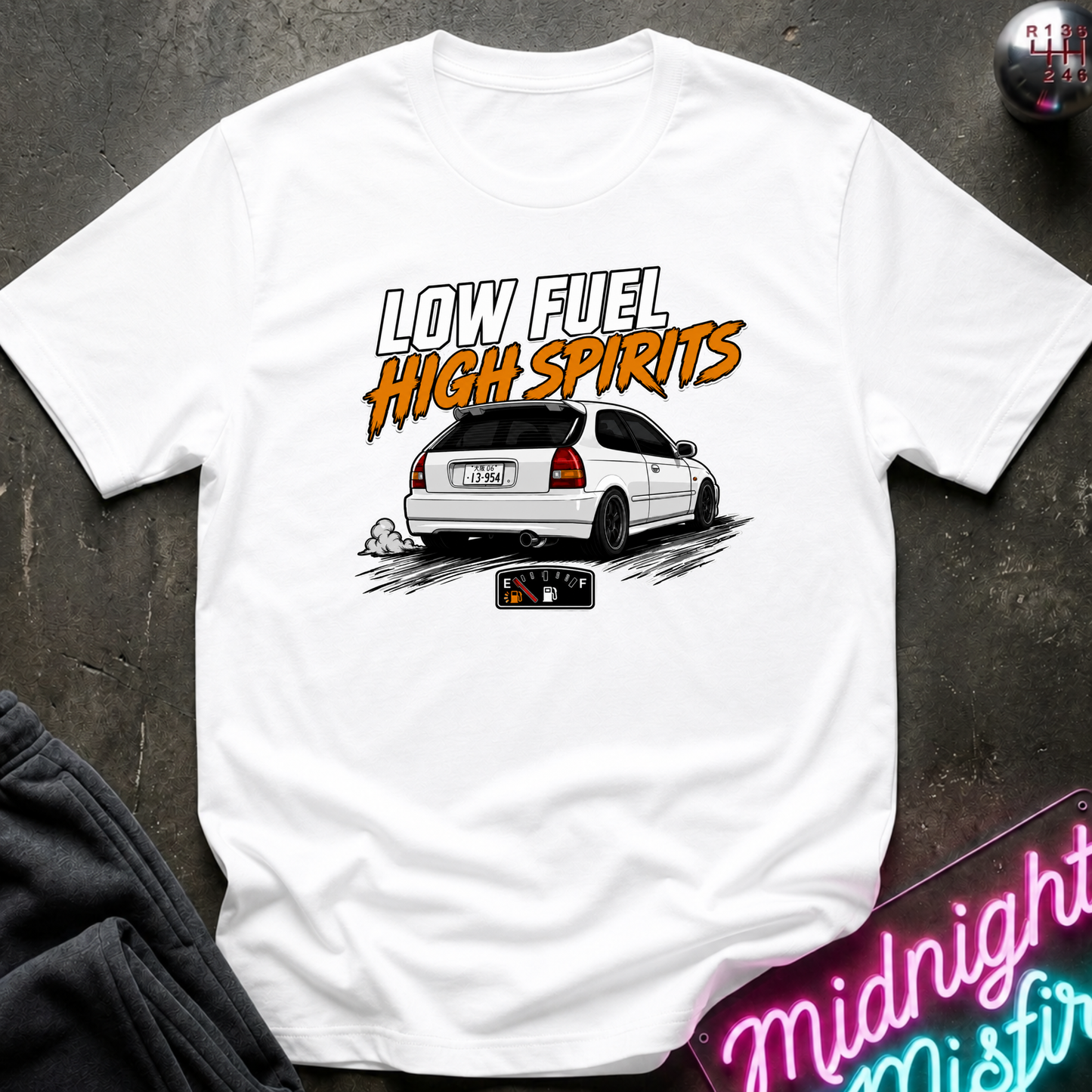 A white short-sleeve graphic t-shirt laid flat on a dark textured surface. The shirt features a graphic print of a white hatchback car with the text 'LOW FUEL HIGH SPIRITS' in stylized orange and white lettering.