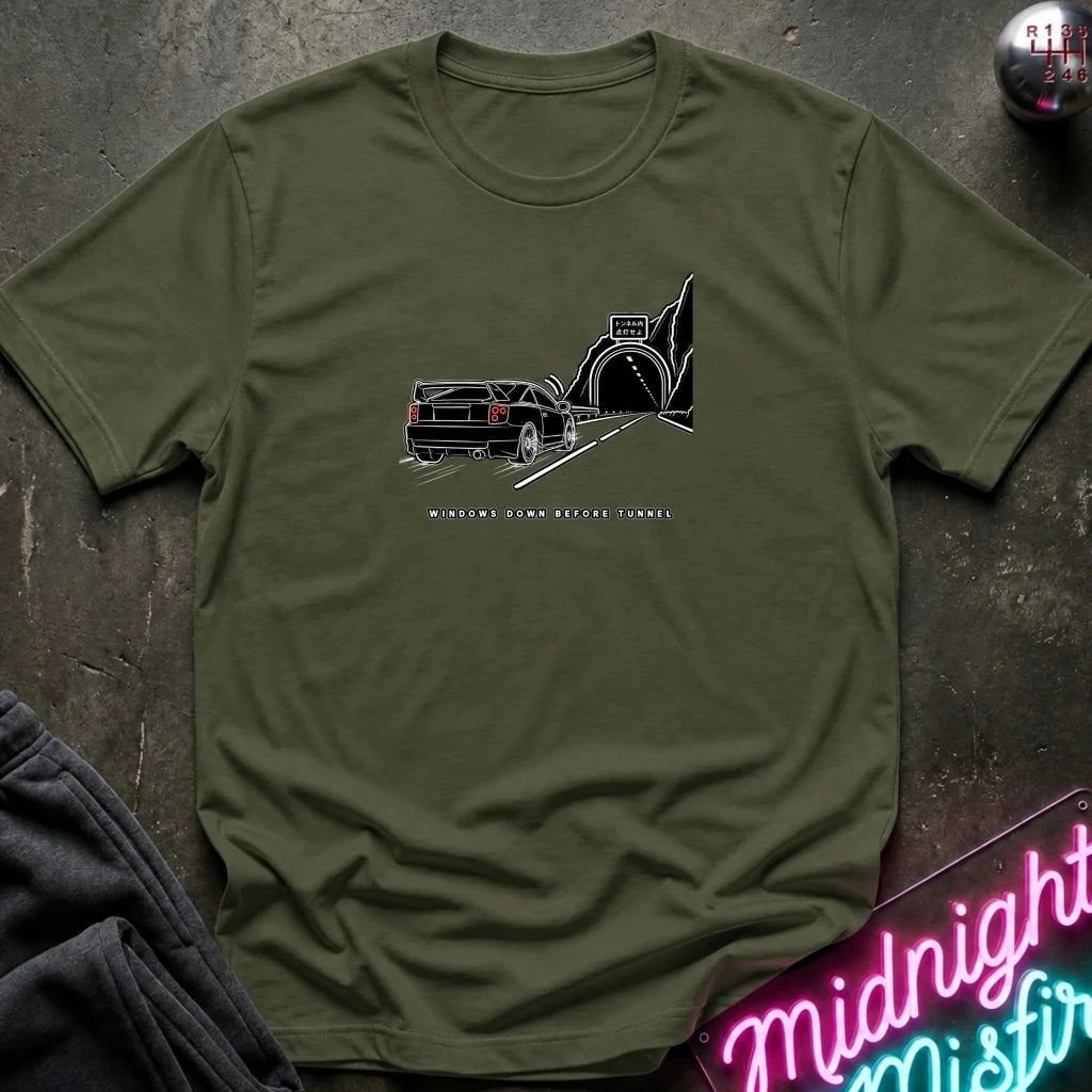 An olive green short-sleeve t-shirt laid flat on a dark textured surface, featuring a black graphic of a classic car in a tunnel with the text.