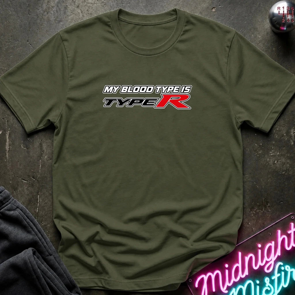 An olive green short-sleeve t-shirt with a 'MY BLOOD TYPE IS TYPE R' graphic printed on the chest, laid flat on a dark textured surface with lifestyle props.