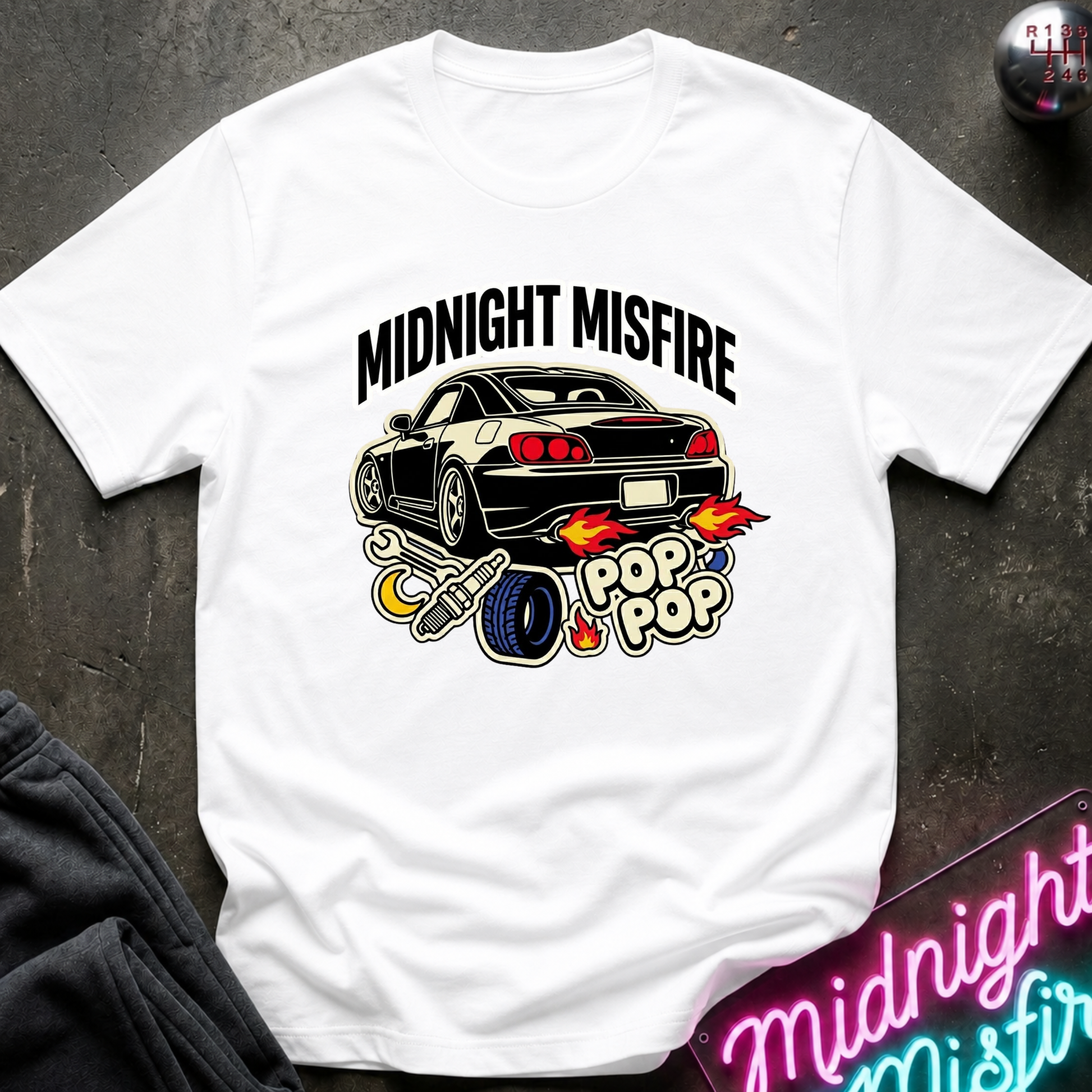 A white short-sleeve graphic T-shirt laid flat on a dark textured surface. The shirt features a black car illustration with flames and the words 'MIDNIGHT MISFIRE' and 'POP POP' in a stylized font.