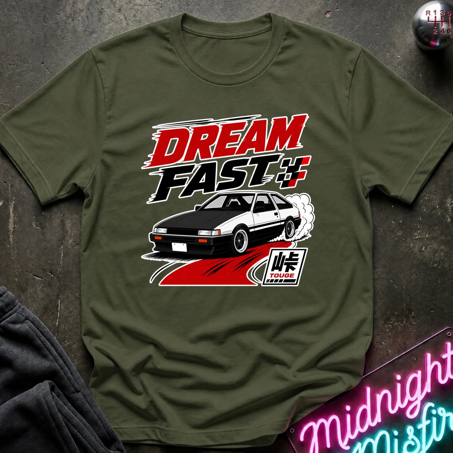 An olive green graphic t-shirt featuring a 'Dream Fast' design with a black and white car, red and white text, and a Japanese flag motif, laid flat on a dark textured surface.