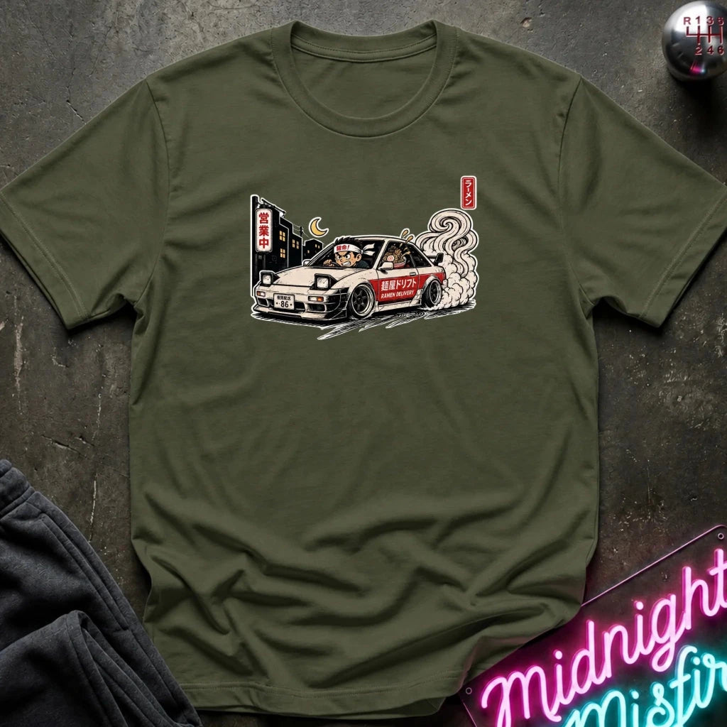 An olive green crewneck t-shirt featuring a graphic print of a white sports car with smoke and Japanese text, styled flat on a dark textured background with a neon sign and dark pants partially visible.