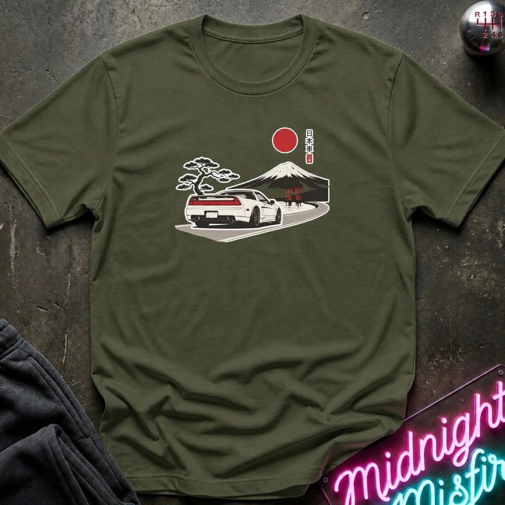 An olive green short-sleeve t-shirt laid flat on a dark textured surface, featuring a graphic print of a white sports car on a road with a red sun, a pine tree, and a mountain in the background. A neon sign and dark clothing are partially visible around the edges.