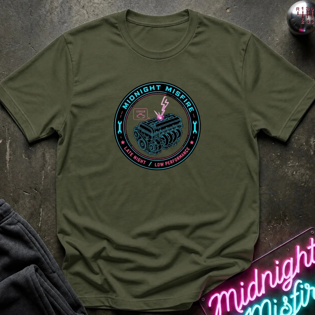 An olive green crewneck t-shirt laid flat on a dark textured surface, featuring a circular graphic print with a tank illustration and the text 'MIDNIGHT MISFIRE'.