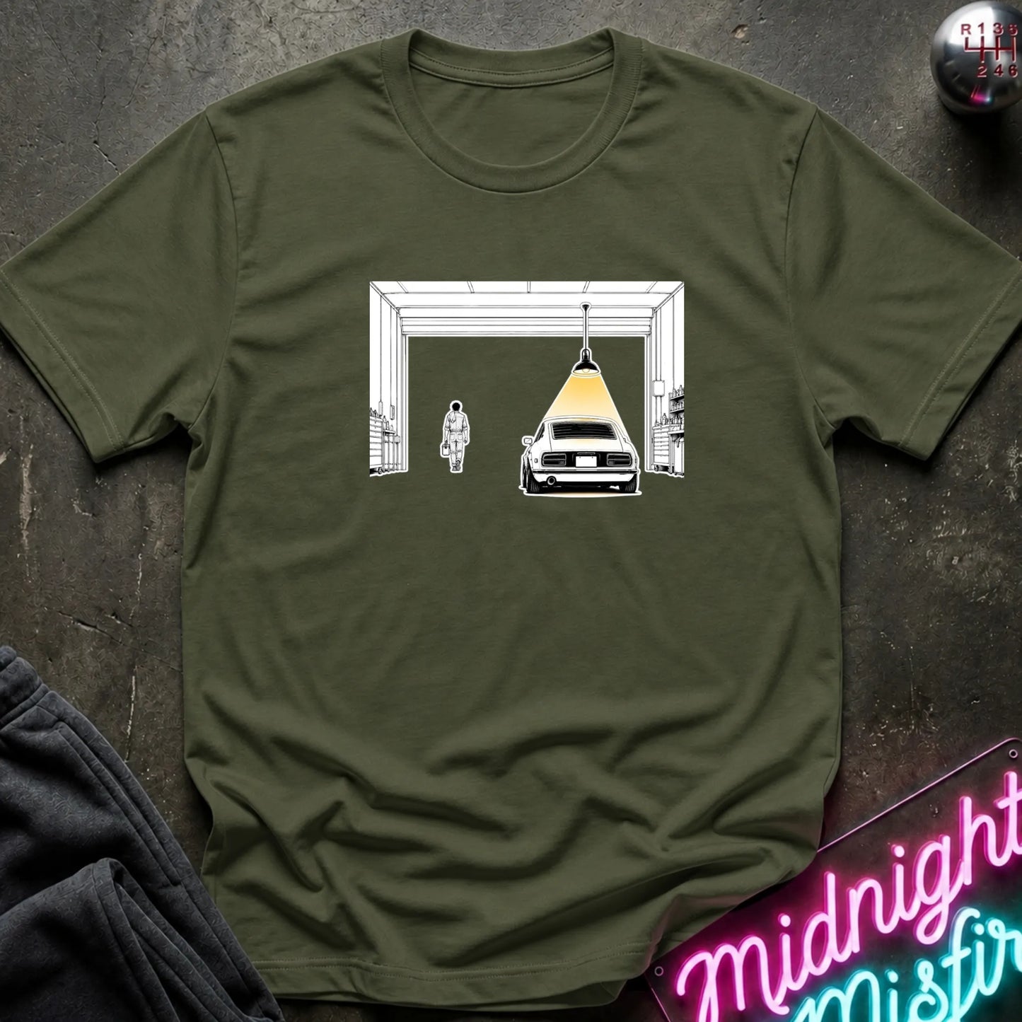 An olive green short-sleeve t-shirt laid flat on a dark textured surface, featuring a graphic print of a car under a hanging lamp inside a garage structure. 