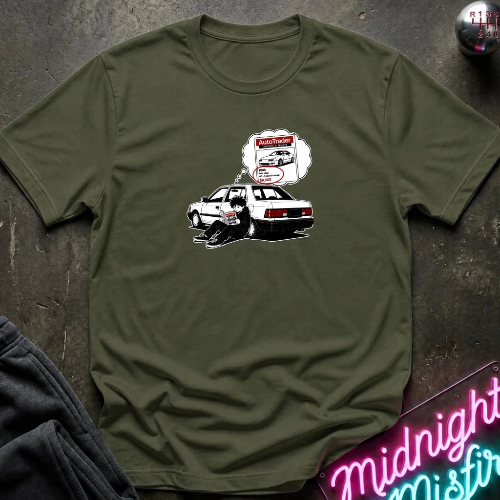 An olive green short-sleeve t-shirt laid flat on a dark textured surface, featuring a graphic of a black-and-whitecar with a thought bubble showing a car part 