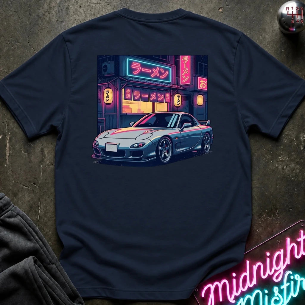 A navy blue short-sleeve t-shirt laid flat on a dark textured surface, featuring a colorful graphic print of a blue sports car parked in front of a neon-lit ramen restaurant. 
