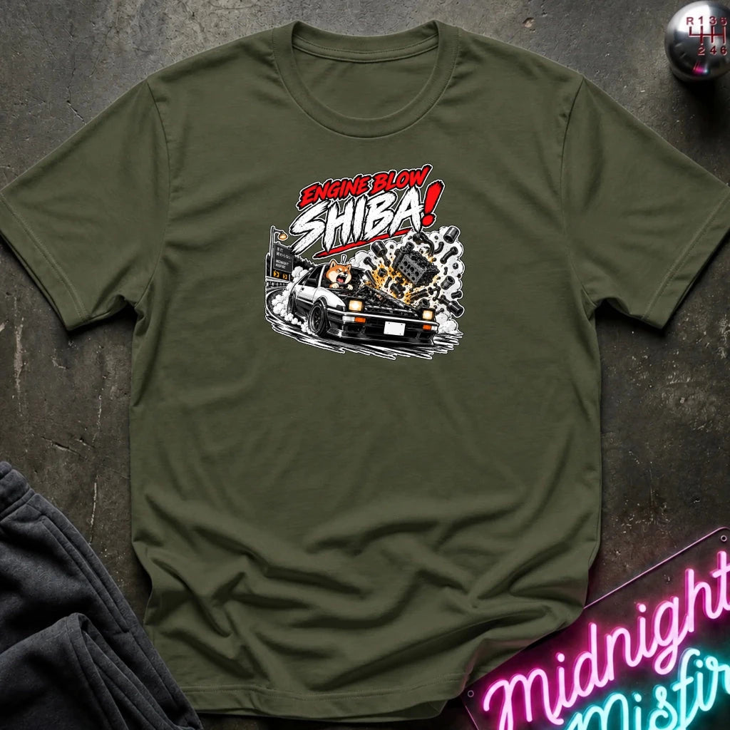 An olive green short-sleeve graphic t-shirt laid flat on a dark textured surface. The shirt features a stylized graphic of a white sports car with a glowing engine and the text 'ENGINE BLOW SHIBA!' in red, white, and black.