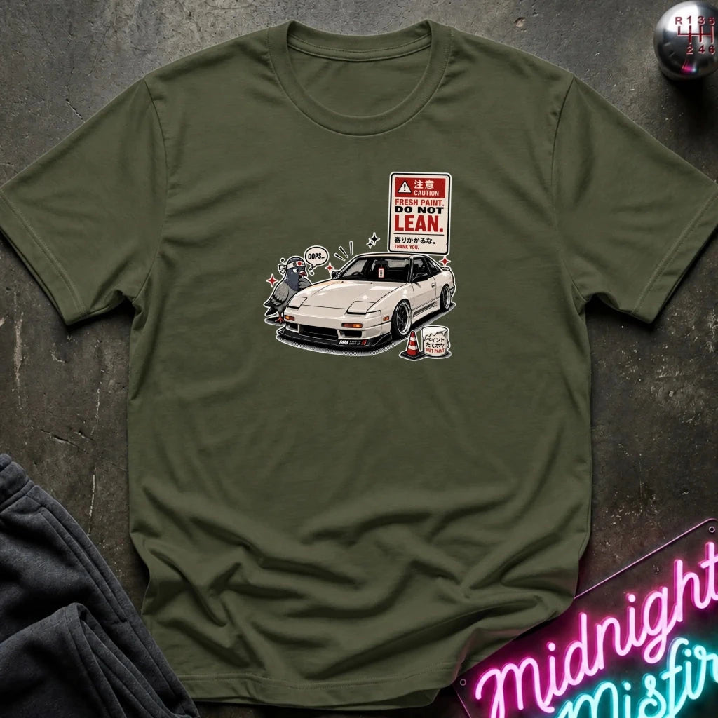 A olive green short-sleeve t-shirt laid flat on a dark textured surface, featuring a graphic print of a white sports car next to a 'DO NOT LEAN' warning sign.